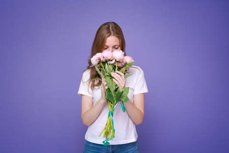 Portrait cheerful young lady with romantic spring bouquet pink peonies on purple studio background. Gorgeous girl enjoys womens day celebration and receives flowers as a special giftの写真素材