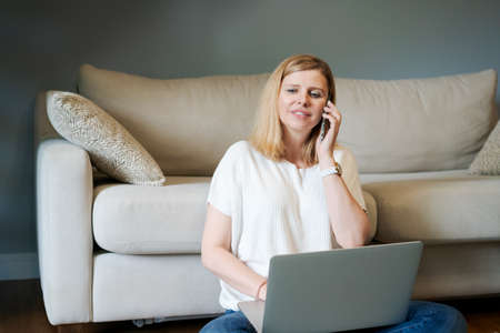 Casual woman, freelancer in blue jeans and light-colored t-shirt, sitting on floor by sofa using...