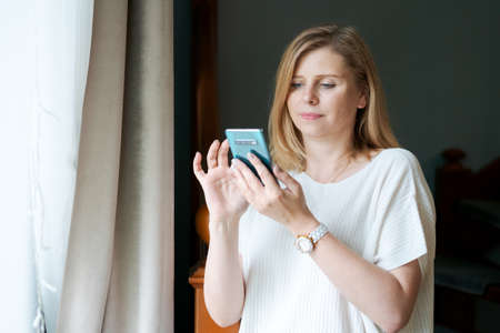 Happy adult woman talking on phone while standing at window against background stairs in living...