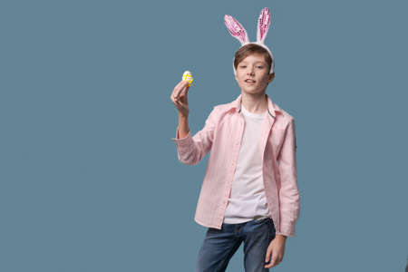 Guy with bunny ears with colorful Easter eggs in his hand in pink shirt. Gesticulating with different funny facial expressions. A boy in a rabbit costume has fun on a blue backgroundの写真素材