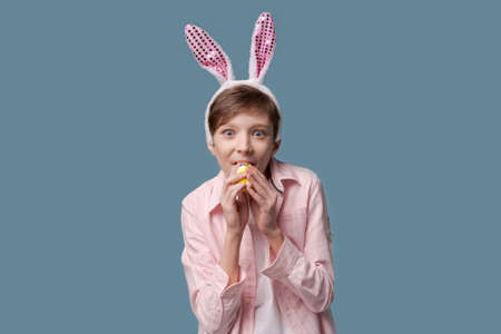 Handsome young man in casual wear pink shirt and bunny easter ears makes different gestures with the palms of his hands with an egg in his hand, looking very funny and funny easter bunny concept.の写真素材