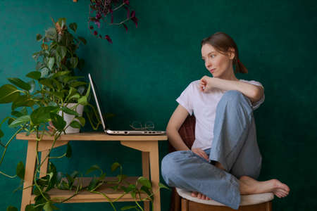 Happy young woman in jeans and white t-shirt at home at table sunny day learning online or remote...
