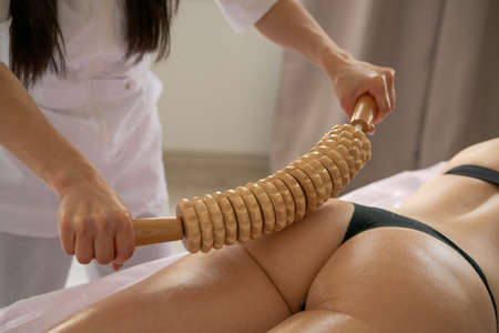 Close-up on back unidentified woman having anti-cellulite massage session with madero therapy, professional therapist holding wooden tools in studio or salon with copy spaceの写真素材