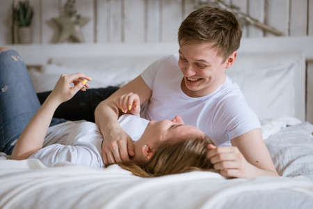 Happy young couple lying on bed smiling as they wake up in trendy hotel room. Wear all white under the covers, spend the day off together, man looking at her in loveの写真素材
