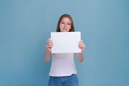 Young woman holding white paper on blue background. Caucasian woman with white blank template sheet with empty space in white t-shirt and blue jeans shows advertisement, mockupの写真素材