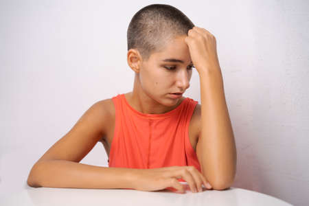 Caucasian girl with short hair, almost bald, holds her hands behind her head, leaning on the table...