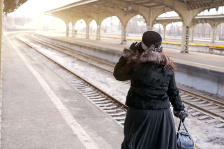 Autumn travel, woman with suitcase is waiting for her train on platform railway station in a black coat and hat. Caucasian lady in warm clothesの写真素材