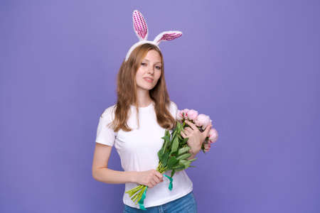 Young smiling woman in pink rabbit ears on her head holds bouquet spring peonies on a purple...