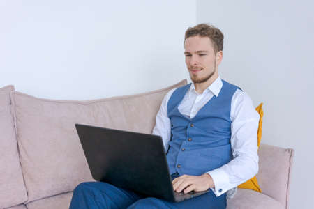 Portrait happy business man with beard in blue suit while sitting in hotel room and using a laptop....