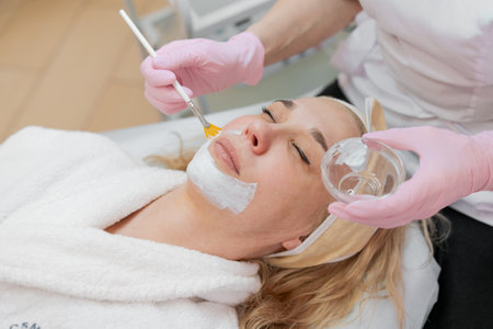 Cosmetology. Professional beautician in medical mask and rubber gloves applies a mask to the clients face. The concept of professional help in beauty salons during a pandemic.の写真素材