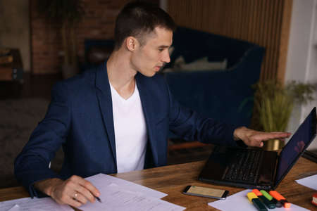 Young man in jacket working from home doing paperwork using laptop and holding a pen in his hand while sitting at his desk. Performs work remotelyの写真素材