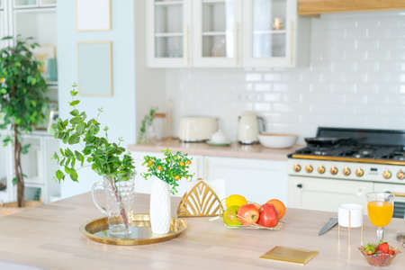 Bright kitchen interior in modern apartment for rent, sale and blog. Kettle and dishes on white furniture, on the table fruit shelf with dishes and a plant in a pot during the daytime, empty spaceの写真素材