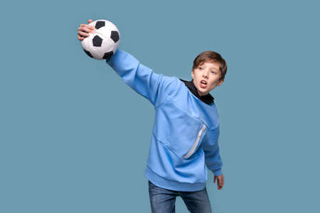 Joyful young man in blue sweatshirt posing isolated on blue wall background. People sincere emotions lifestyle concept. Layout copy space. Holding soccer ball, making winner gesture, yelling teenagerの写真素材