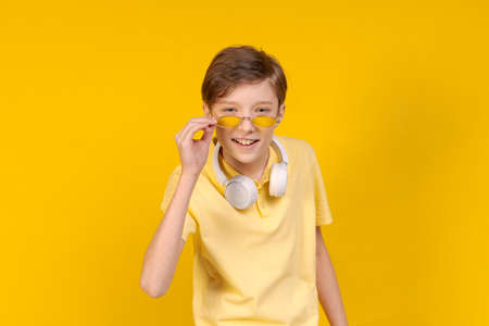 Studio happy smiling young male wearing bright t-shirt and round glasses, with headphones around his neck, listening to his favorite music, looking in on yellow background. Caucasian teenager posingの写真素材