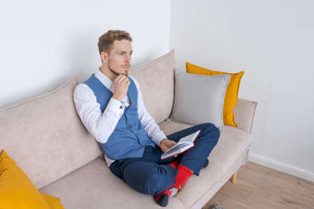 Pensive businessman sits on sofa with diary in his hand, thinks about plans in a blue bearded guy suit. Plans his daily routine and business meetings. Record keeping and planning conceptの写真素材