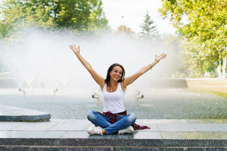 Cheerful young brunette woman against background fountain happy shows gesture with her hands from happiness. Caucasian girl on sunny day against background water with rainbow rejoices in carefree lifeの写真素材