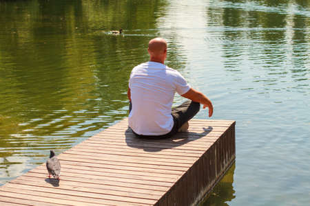 Lonely man sitting on edge wooden raft on pier looks out over lake on sunny day in the park. Solo travel through nature, wanderlust and inspirational conceptの写真素材