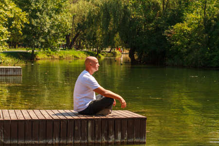 Lonely man sitting on edge wooden raft on pier looks out over lake on sunny day in the park. Solo travel through nature, wanderlust and inspirational conceptの写真素材