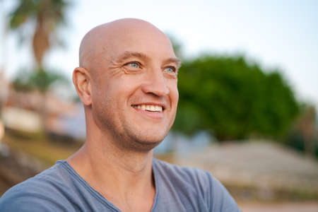 Male portrait happy man with bald head in blue t-shirt smiles with his teeth and looks away in a blue t-shirt against the backdrop of trees and sky on the seashoreの写真素材