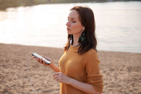 Woman with white headphones relaxing on lake and listening to music at sunset. Young beautiful caucasian girl listens to music with headphones in nature - music, relax calmness on a summer eveningの写真素材