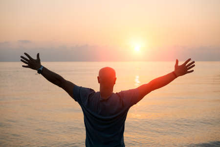 Silhouette man with his hands raised at sunset on beach arms out to sides towards the sun, concept for religion, worship, prayer and praise and success in life, peace and tranquility in natureの写真素材
