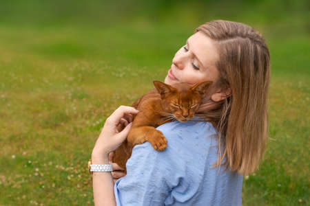 Satisfied woman with her pet ginger cat sitting in an armchair on grass on beautiful meadow, afternoon in the yard at the cottage, Woman stroking her beloved pet holding him in her armsの写真素材