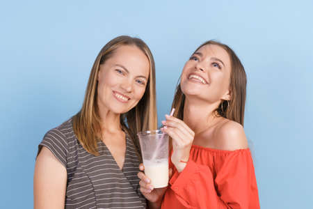 Happy mom and adult daughter drink milkshakes and laugh merrily in dresses on a blue background in...