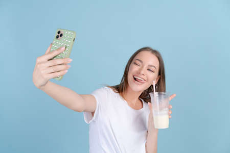 Happy beautiful girl takes picture herself with fresh milkshake on mobile phone in white t-shirt or talks with friends via video call. Caucasian young woman smiling with teeth, Blue Studio backgroundの写真素材