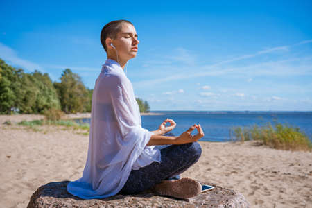 Graceful woman doing yoga asana on beach in headphones meditates on seashore on a sunny day. Caucasian girl in sportswear sits in the lotus position and relaxesの写真素材