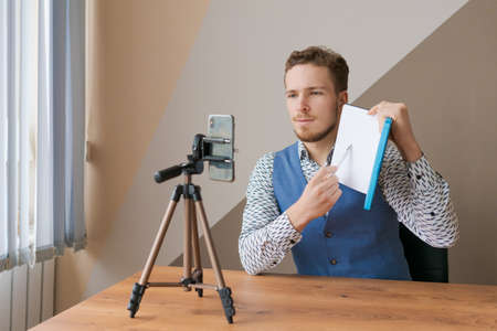 Smiling business man using phone and tripod, making video call for co-workers, sitting in office at...