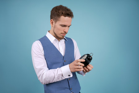 Young business man stands with an alarm clock in his hands on a blue studio background. The concept...