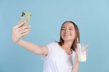 Happy beautiful girl takes picture herself with fresh milkshake on mobile phone in white t-shirt or talks with friends via video call. Caucasian young woman smiling with teeth, Blue Studio backgroundの写真素材