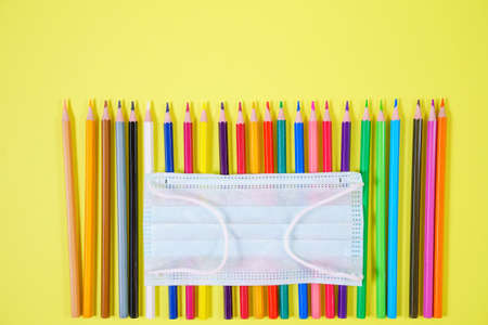 Multicolored colored pencils close-up from above protective mask on yellow background, school time quarantine conceptの写真素材