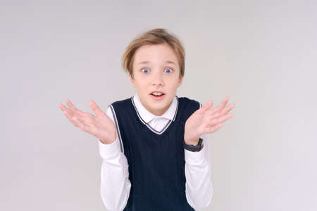Photo an adorable young happy boy looking at camera and gesturing with his hands emotionally in a white shirt and blue tank top in a school uniform against a light background.の写真素材