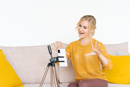 Cheerful caucasian woman sitting on sofa using mobile phone talking on video link and gesturing,...