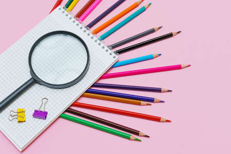 Magnifying glass, pencils and a blank white notepad on a spring on a pink background. Back to school...