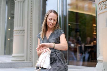 Street style cute woman blogger in gray dress and analog wrists. Stylish woman checks the time on her watch. Sits on a bench near the store with a handbagの写真素材