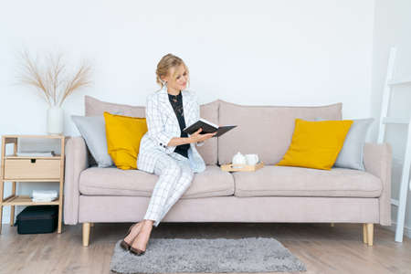 Middle age woman working at home in business suit sitting on couch using reading notebook looks at...