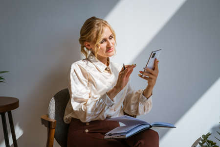Attractive woman in business clothes with a smartphone in her hands, next to a table in an office...
