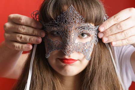 Male hands put lace mask on face beautiful girl with red lipstick on red background close-up, looking at the camera young caucasian womanの写真素材