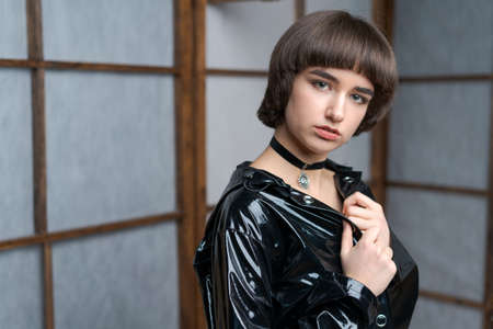 Portrait beautiful young caucasian woman in black patent suit with one hand holding pistol to her head, young sexy short hair girl with pistol looking at camera, pretty women are standing with pistolの写真素材