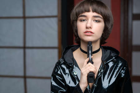 Portrait beautiful young caucasian woman in black patent suit with one hand holding pistol to her head, young sexy short hair girl with pistol looking at camera, pretty women are standing with pistolの写真素材