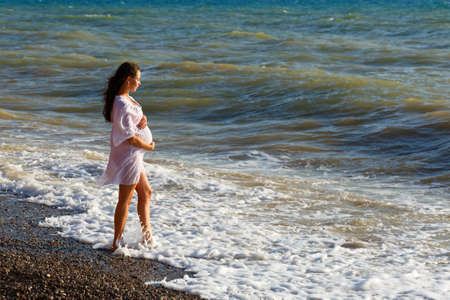 Pregnant woman stands on sea beach in summer in white light dress with fluffy hair in the wind. Enjoys the wind and brags from the waves. Waiting for a baby, happy young womanの写真素材