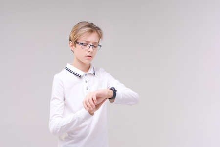 A happy boy in glasses and a shirt looks at his watch or fitness tracker. Isolated on a gray background. Schoolboy. Teenager.の写真素材