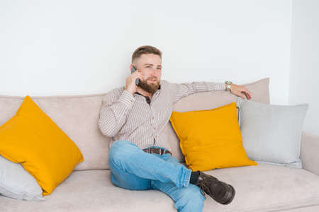 Portrait an attractive smiling young bearded man dressed in casual clothes shirt and jeans sitting on a sofa in the living room talking on a mobile phoneの写真素材