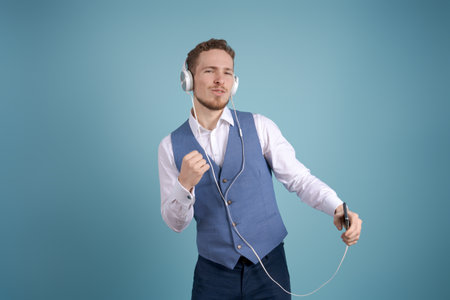 Technology, gadgets and people. Carefree business man listening to his favorite song in headphones, using a mobile phone, singing and dancing on a blue backgroundの写真素材