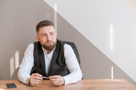 Casual business man bearded, sitting at table with pen in his hand, thinking about a work project, dressed in a shirt in the office. Startup business, portable office conceptの写真素材