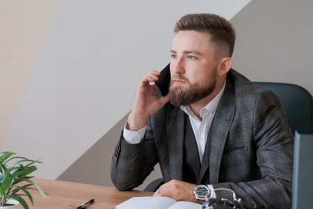 Focused bearded business guy in office clothes sitting at table and talking on smartphone, noting...