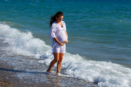 Beautiful pregnant woman standing on beach in light dress in the wind. Pleasure of waiting for a child. Happy young caucasian woman with flowing long hairの写真素材