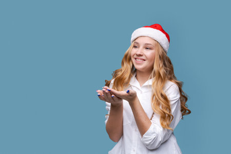 Cute happy girl in santa claus hat holding hand waiting for gift, in white shirt on blue background in the studio, copy space, concept of winter sales for christmas and new yearの写真素材